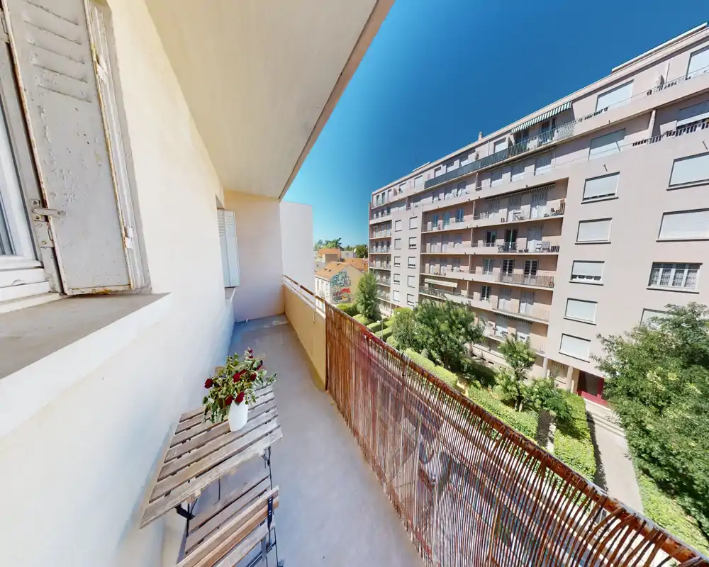 Balcon privé ensoleillé avec sol étroit, claustra en bambou et une petite table fleurie, donnant sur une cour calme et un immeuble voisin.
