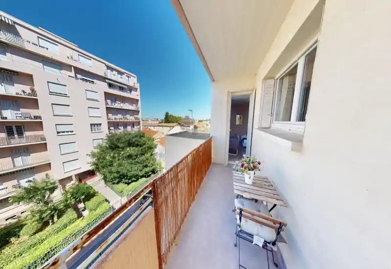 Balcon ensoleillé avec petite table et deux chaises, donnant sur un immeuble résidentiel et une cour arborée — idéal pour le café du matin.