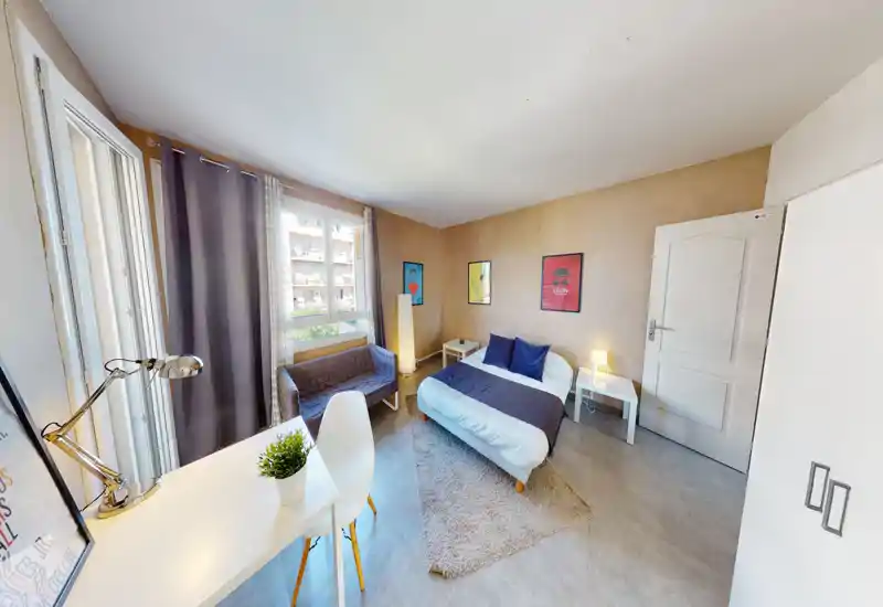 Chambre lumineuse et bien mise en scène avec lit double, tables de chevet et tapis douillet. Grande fenêtre apportant lumière naturelle ; bureau et coin salon pour plus de fonctionnalité.