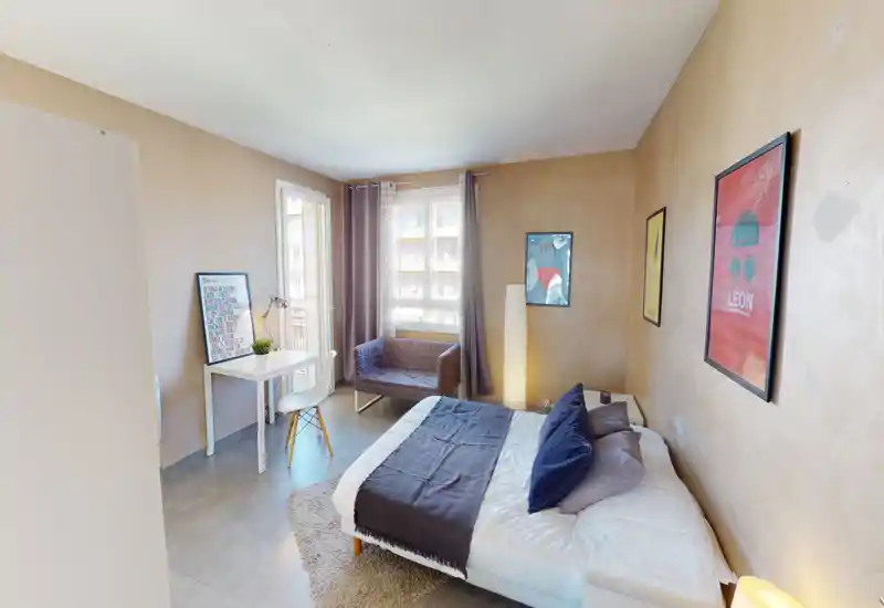 Chambre lumineuse et élégante avec grand lit en linge sombre, petit bureau près de la fenêtre et fauteuil confortable — idéale pour un séjour agréable et du télétravail.