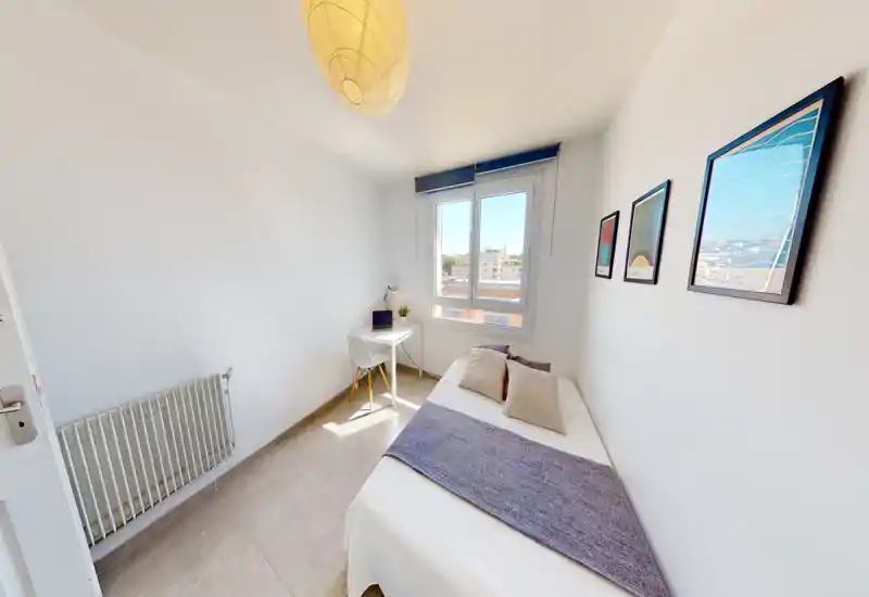 Chambre compacte et ensoleillée avec lit simple, coussins décoratifs et un petit bureau près de la fenêtre apportant une belle lumière naturelle.