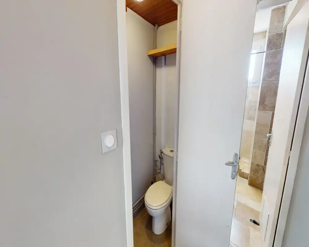 Salle de bain compacte avec WC individuel, carrelage neutre et plomberie apparente ; espace petit mais fonctionnel.