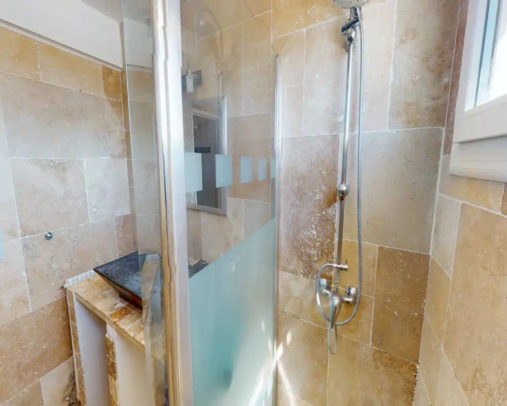 Salle de bain carrelée et lumineuse avec douche vitrée, mitigeur mural et douchette; la lumière naturelle provenant de la fenêtre éclaire l'espace.