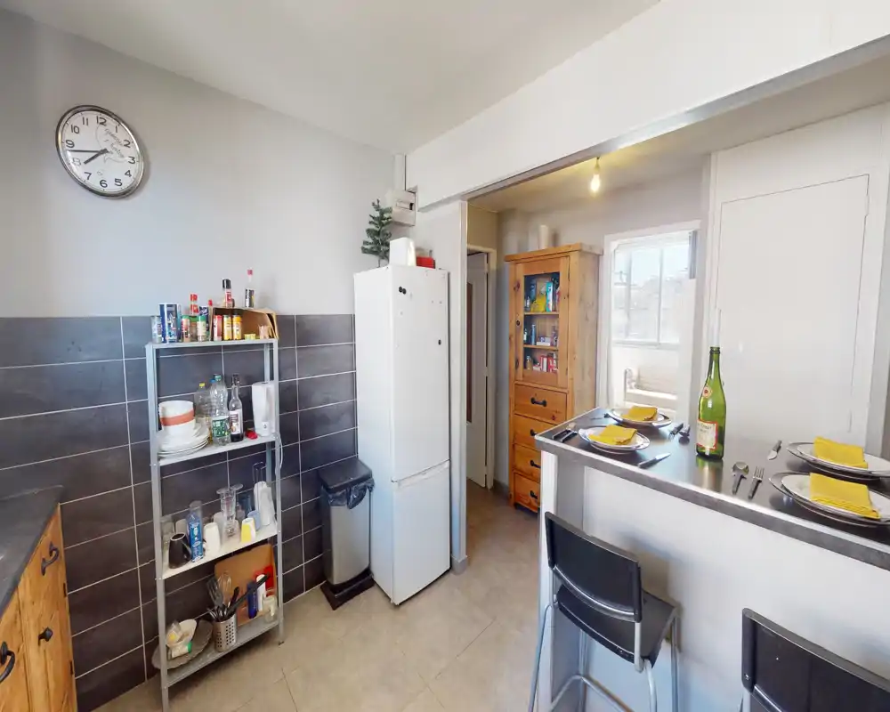Cuisine lumineuse et fonctionnelle avec réfrigérateur, étagères ouvertes, meubles en bois et bar pour le petit-déjeuner pour deux — soignée et pratique pour de courts séjours.