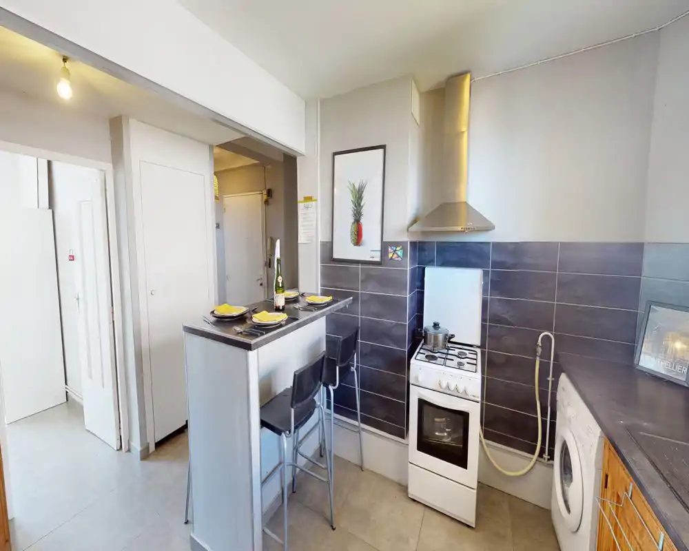Cuisine compacte et bien mise en scène avec bar pour le petit-déjeuner, cuisinière et lave-linge. Carrelage neutre et petit comptoir pour un espace de cuisson lumineux et fonctionnel.