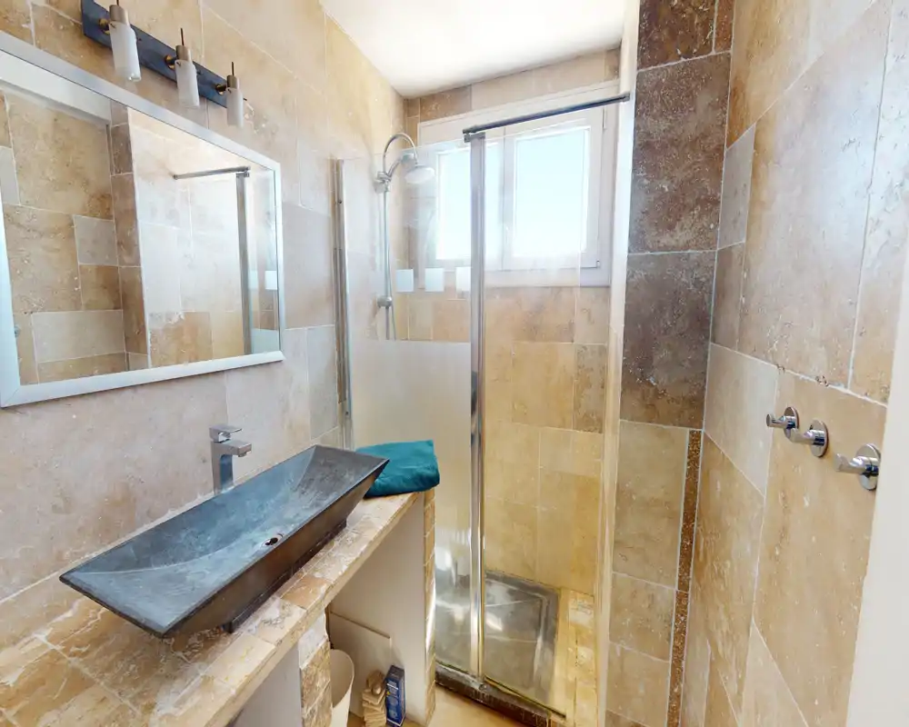 Salle de bains lumineuse avec carrelage en pierre, douche, vasque contemporaine, miroir et robinetterie chromée — compacte et bien entretenue.