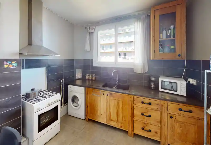 Cuisine lumineuse et fonctionnelle avec des placards en bois, une cuisinière à gaz, un évier sous la fenêtre et un lave-linge compact — espace chaleureux et pratique.