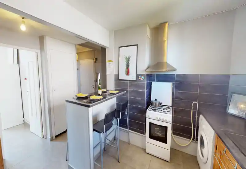 Cuisine compacte et bien mise en scène avec bar pour le petit-déjeuner, cuisinière et lave-linge. Carrelage neutre et petit comptoir pour un espace de cuisson lumineux et fonctionnel.