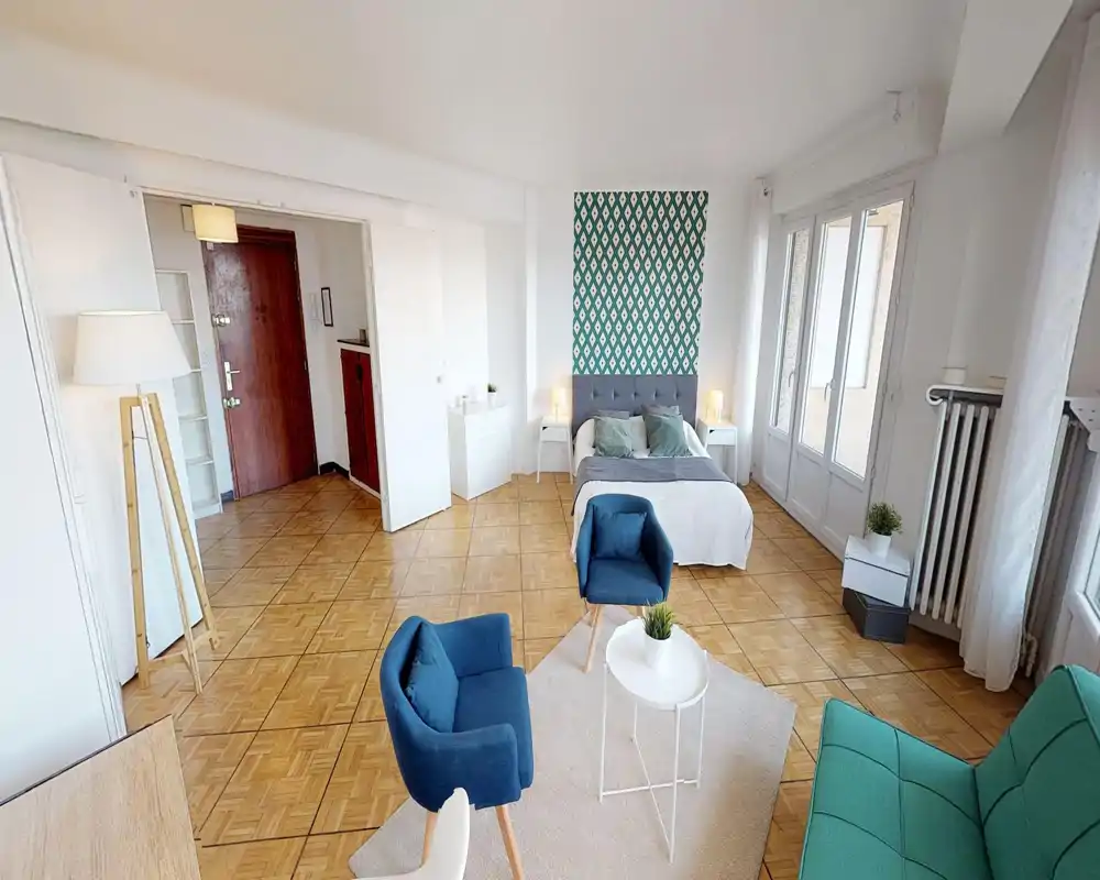 Chambre/studio lumineuse et bien mise en scène avec un lit double devant un mur décoratif, tables de chevet avec lampes, coin salon avec fauteuils bleus et petite table basse, et de grandes fenêtres apportant beaucoup de lumière.