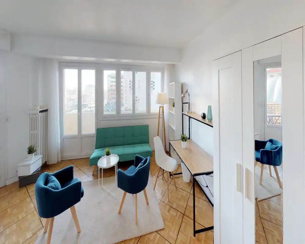 Salon moderne et lumineux avec un canapé bleu-vert, deux fauteuils bleus et un espace de travail le long du mur. De grandes fenêtres apportent une belle lumière naturelle et le parquet clair réchauffe la pièce.