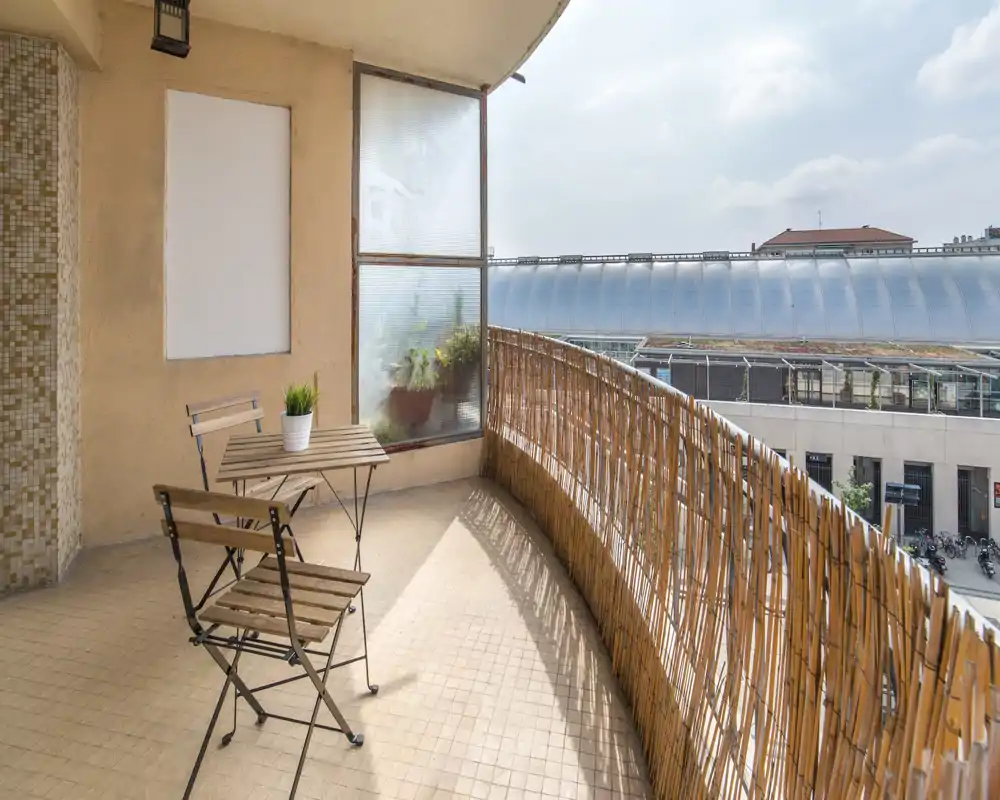 Balcon ensoleillé avec petite table bistrot en bois et deux chaises pliantes, brise-vue en bambou et vue urbaine — idéal pour un café matinal ou un moment de détente en plein air.
