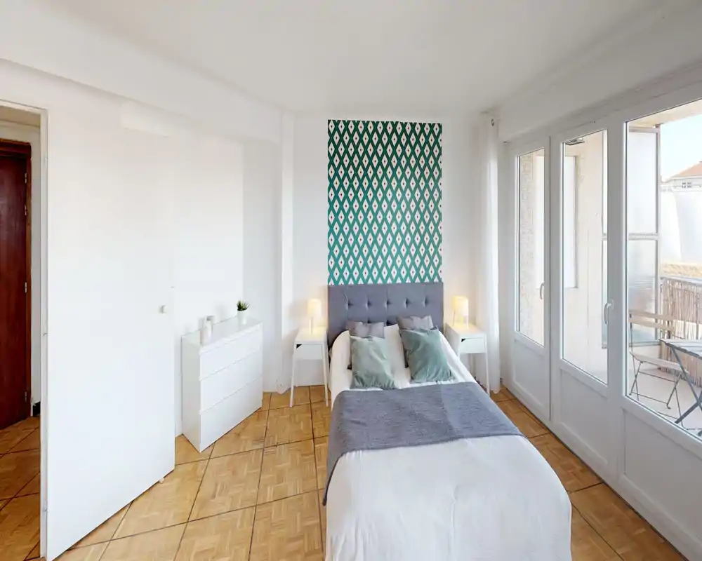 Chambre lumineuse et moderne avec tête de lit capitonnée, mur d'accent à motif et grandes fenêtres donnant sur un balcon. Linge de lit blanc propre, tables de chevet avec lampes et coussins décoratifs créent une atmosphère chaleureuse et lumineuse parfaite pour une annonce.
