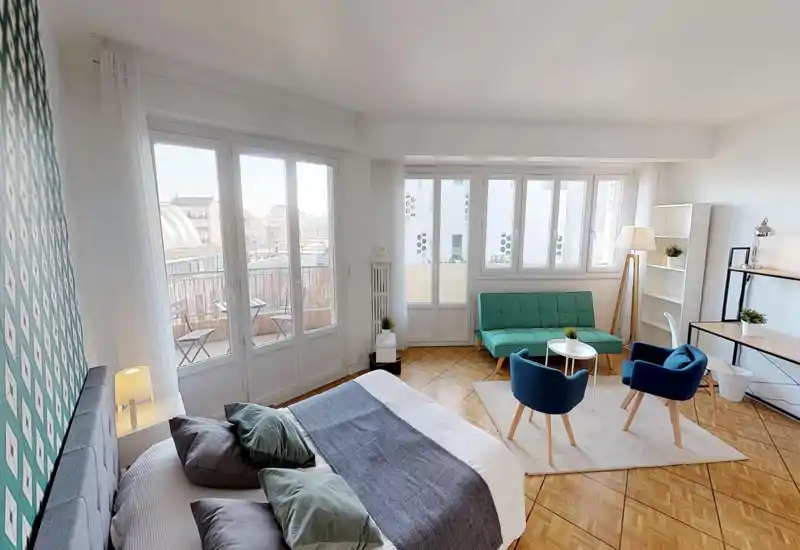 Chambre-studio spacieuse et lumineuse avec un lit double bien fait au premier plan, de grandes fenêtres et un coin salon cosy avec canapé et fauteuils — idéale pour annonces immobilières.