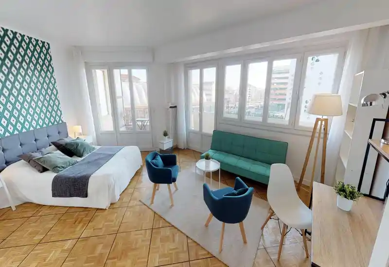 Chambre-studio spacieuse et lumineuse avec un grand lit confortable, un coin salon et de grandes fenêtres qui laissent entrer beaucoup de lumière naturelle. La pièce dispose d'un mur d'accent à motifs, de textiles douillets et d'un petit salon avec canapé et fauteuils.