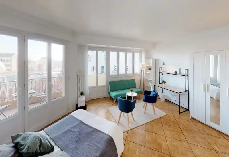 Chambre moderne et lumineuse avec de grandes fenêtres et un lit double confortable au premier plan, accompagnée d'un coin salon avec canapé et fauteuils — idéale pour un appartement meublé en ville.