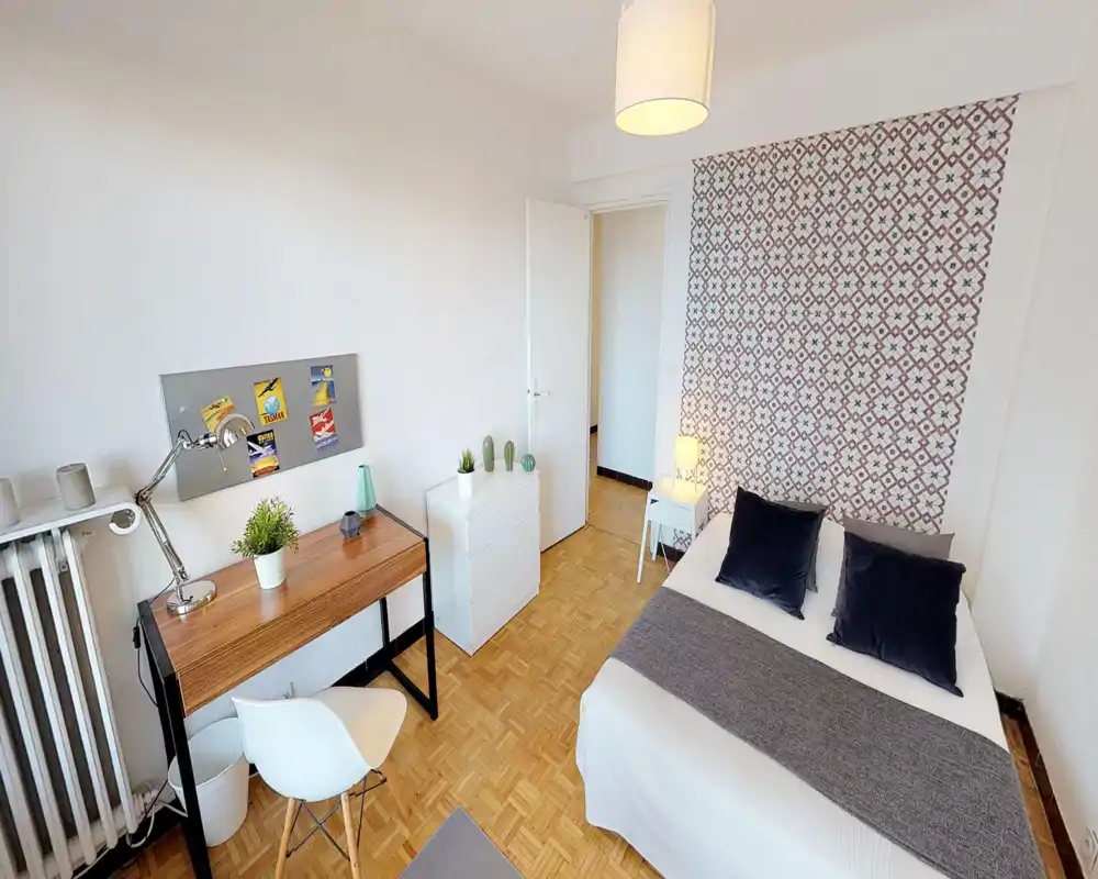 Camera da letto luminosa e curata con letto matrimoniale, parete decorativa a motivi, comodino con lampada e una scrivania per lavorare. Perfetta per chi cerca riposo e un’area studio.