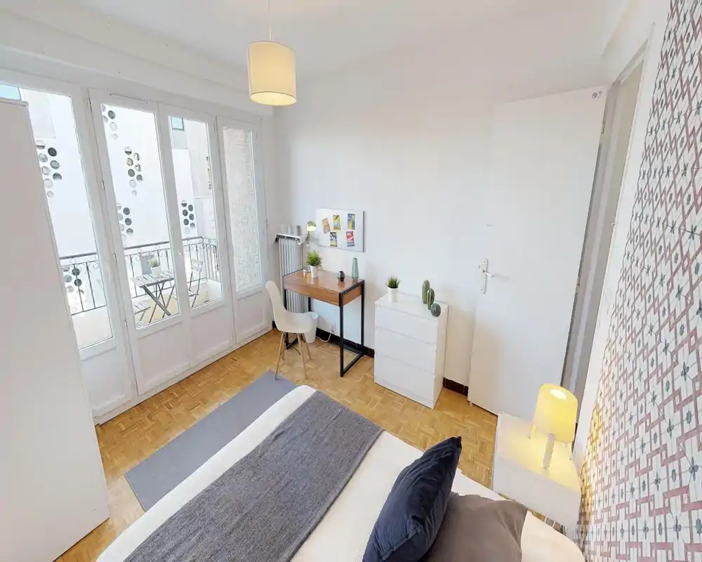 Camera da letto luminosa e moderna con un ampio letto in primo piano, una scrivania e un comò vicino alla parete e finestre a tutta altezza che danno su un balcone — ariosa e ben illuminata.