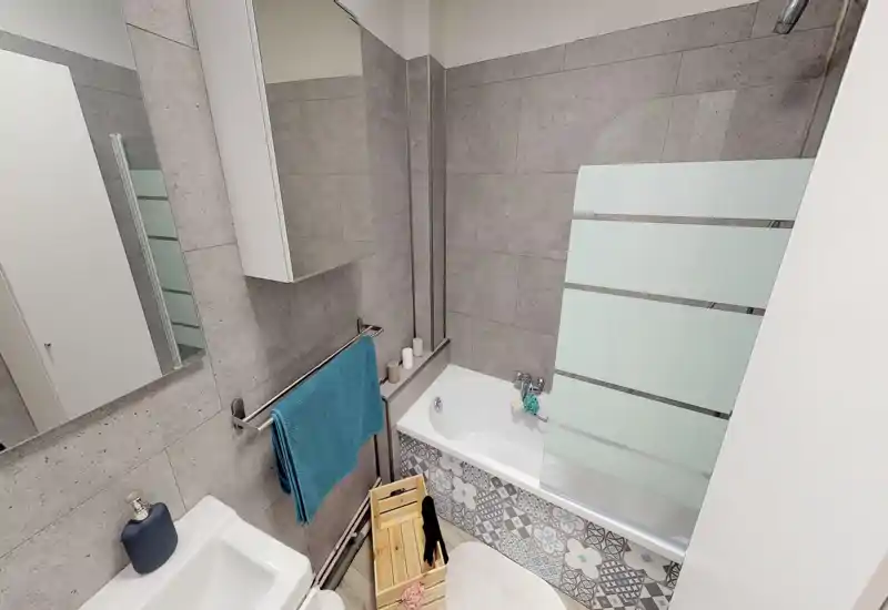 Salle de bains moderne et compacte avec baignoire/douche, carreaux à motifs et revêtement mural gris. Une serviette, des articles de toilette et une petite caisse en bois apportent une touche pratique.