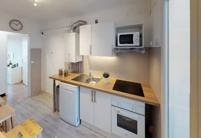 Cuisine compacte et moderne avec plans de travail en bois, four encastré et plaque à induction, micro-ondes et lave-vaisselle. Carrelage clair et éclairage sous meuble offrent un espace de travail lumineux et pratique.