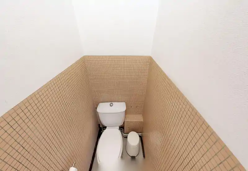 Petite salle de bain avec WC à réservoir, carreaux carrés beiges couvrant la moitié inférieure des murs et murs supérieurs peints en blanc. Espace réduit et fonctionnel avec une petite poubelle et plomberie apparente au sol.