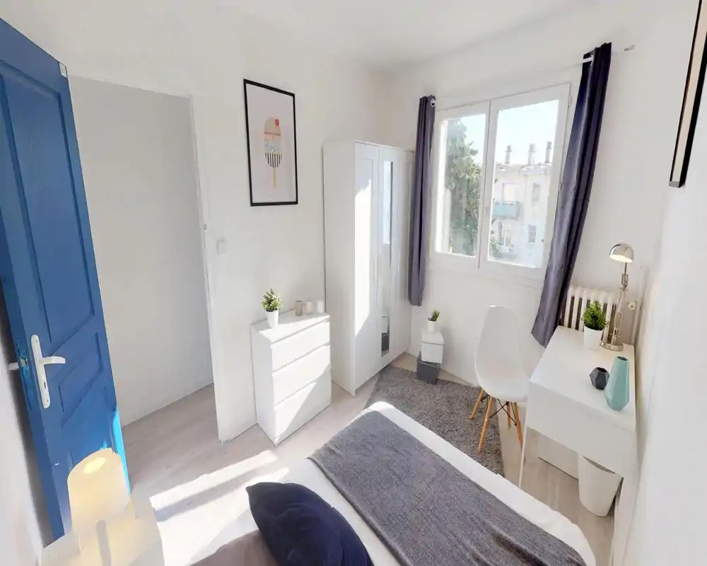 Chambre lumineuse et soignée avec lit au premier plan, mobilier blanc, armoire, bureau et lumière naturelle provenant d'une grande fenêtre.