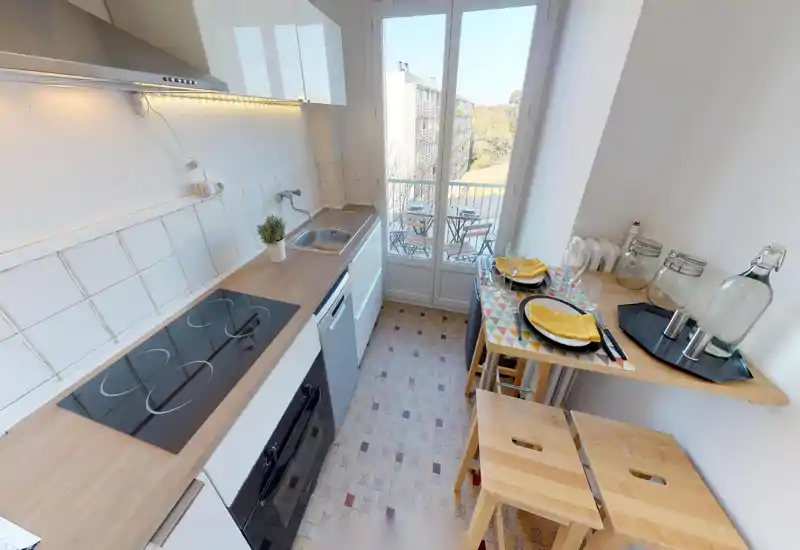 Cuisine compacte et lumineuse avec plaque à induction moderne, évier et petit bar donnant sur un balcon ensoleillé — mise en scène soignée avec vaisselle et bocaux en verre.