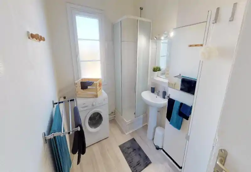 Salle de bains lumineuse et soignée avec douche d'angle, lavabo sur pied et machine à laver ; sol effet bois et serviettes bleues pour une ambiance fraîche et propre.