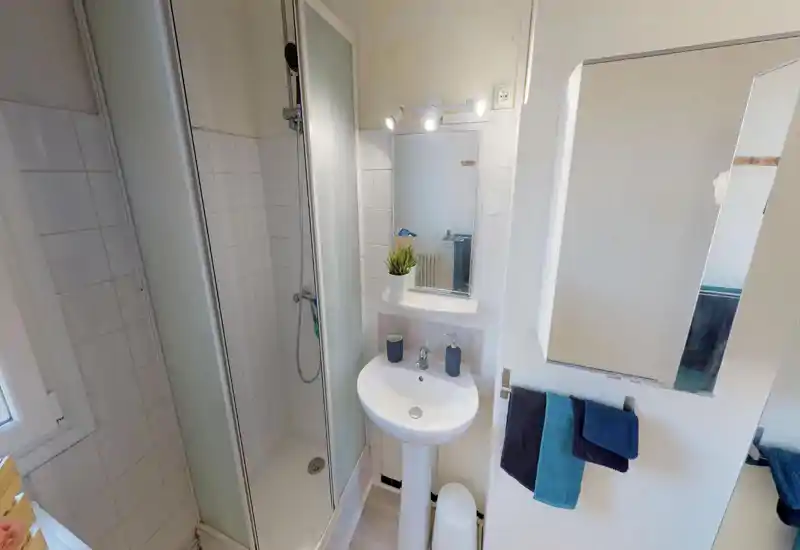 Salle de bains compacte et propre avec douche d'angle, lavabo sur colonne et meuble miroir. Lumière naturelle et équipements simples et modernes pour un usage pratique.