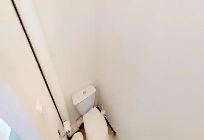 WC compact avec cuvette standard dans un petit coin bien éclairé ; brosse de toilette et porte-papier mural visibles.