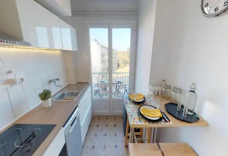 Cuisine compacte et lumineuse avec plan de travail en bois, plaque à induction, évier et petite table pour deux près d'une porte-fenêtre laissant entrer la lumière naturelle.