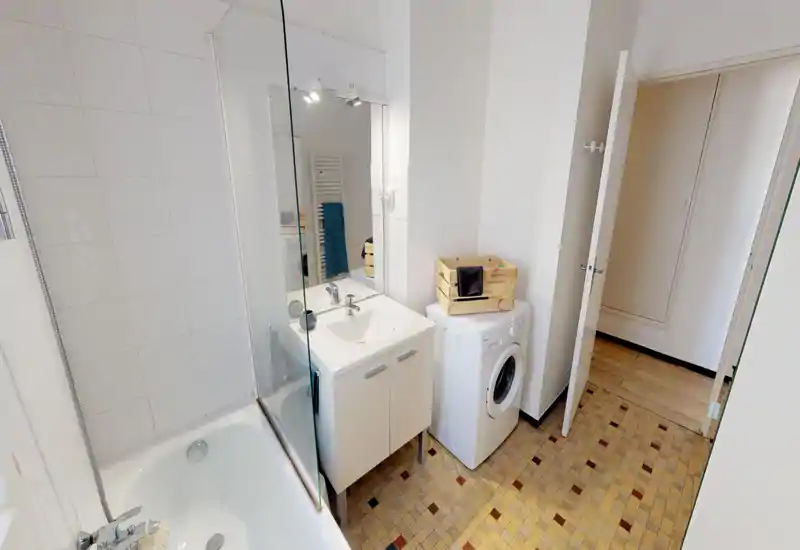 Salle de bain lumineuse et soignée avec baignoire-douche, lavabo avec meuble et machine à laver. Carrelage neutre et lumière agréable pour un espace propre et fonctionnel.