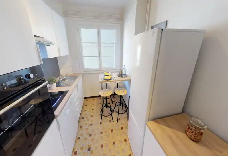 Cuisine compacte et lumineuse avec des meubles blancs modernes, un four encastré et un petit bar avec deux tabourets. Le sol carrelé et la décoration simple offrent un espace fonctionnel et agréable.