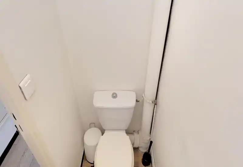 Toilettes compactes avec réservoir blanc, plomberie apparente et balayette ; petit WC propre et fonctionnel, adapté pour une annonce locative.