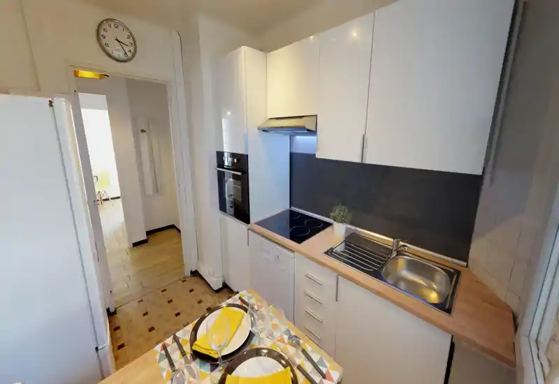 Cuisine moderne compacte avec placards blancs, plaque à induction, four encastré et évier en acier. Un petit coin repas avec serviettes jaunes et une plante apporte une touche chaleureuse.