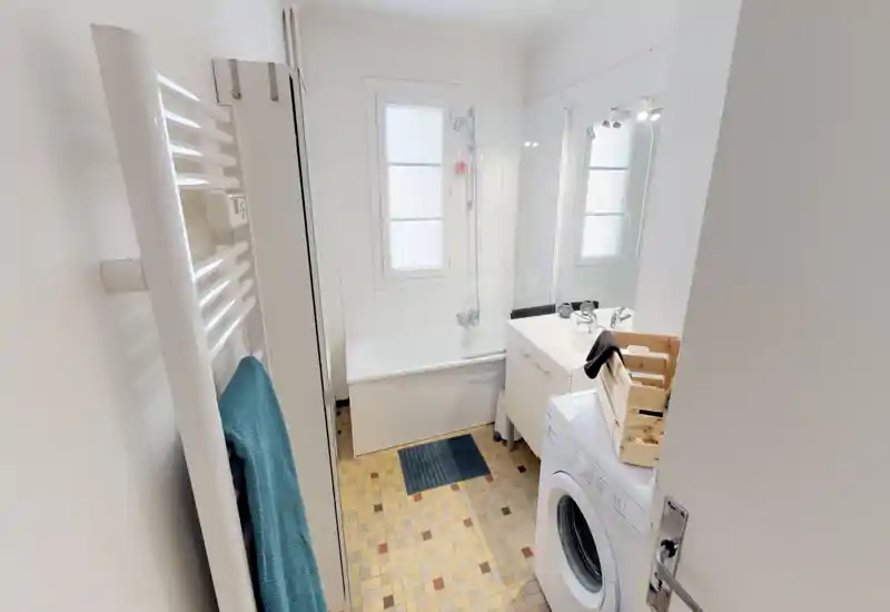 Salle de bains lumineuse et propre avec baignoire/douche, lavabo avec meuble et lave-linge; sol en mosaïque et porte-serviettes chauffant pour plus de confort.