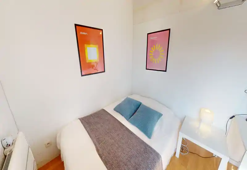 Small, cozy bedroom with a double bed dressed in white linens, two blue cushions and a textured runner; simple bedside table with lamp and framed artwork above the bed.