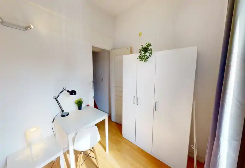 Compact bedroom with a white wardrobe and a small desk by the window. Neutral, minimal décor with natural light and simple workspace—ideal for a single occupant or student.
