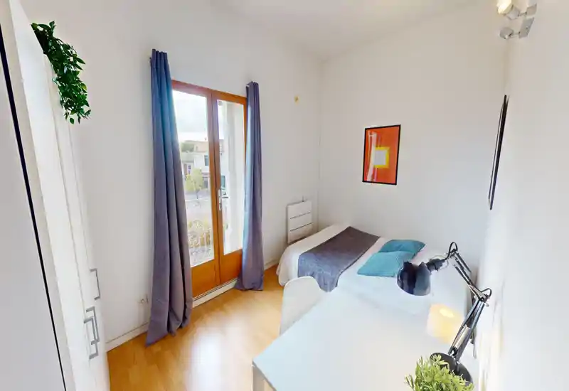 Bright single bedroom with a small desk and natural light from a balcony door. The room is neatly furnished with cozy textiles and a modern desk lamp, ideal for a student or solo traveler.
