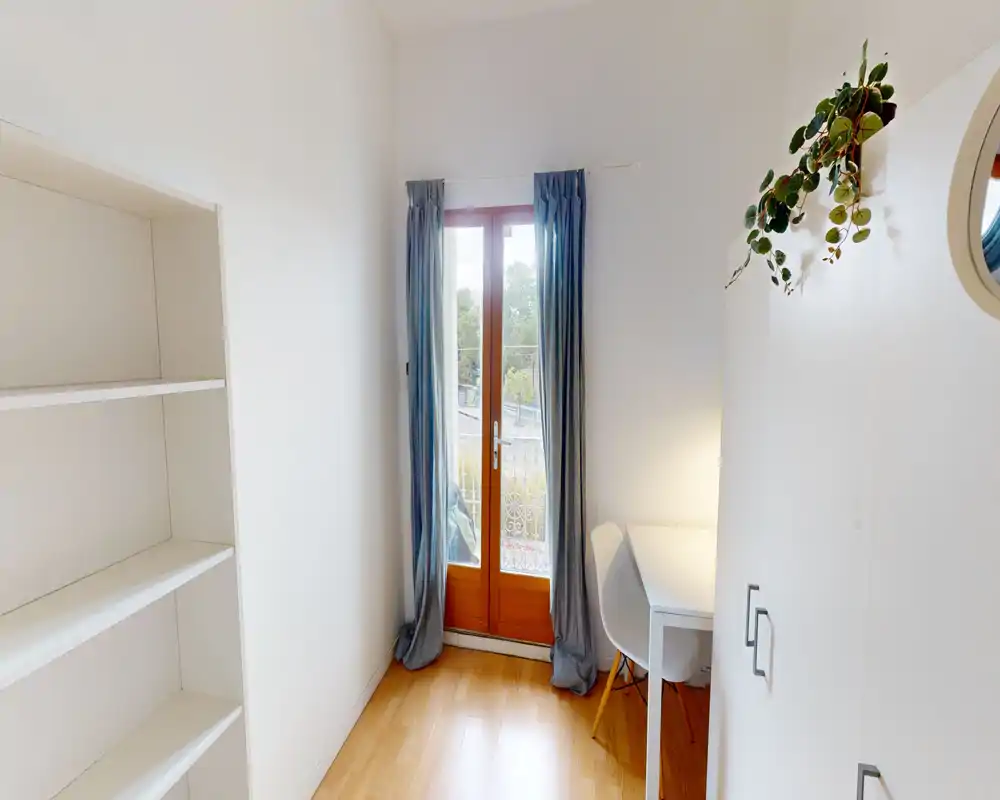 Compact bright living area with a glass door opening to a balcony, soft wood floors, a small desk and cozy curtains—ideal for a studio setup.