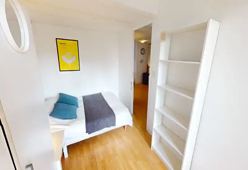Compact, bright bedroom with a neatly made double bed, decorative cushions and a simple wall print; light wood flooring and a tall white shelving unit add a clean, modern look.