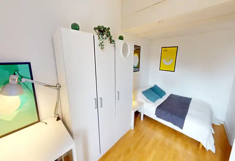 Neat single bedroom with a white wardrobe, bedside table and a well-made single bed with teal cushions and a decorative throw. Bright, compact and simply furnished — ideal for a solo tenant or short-term stays.