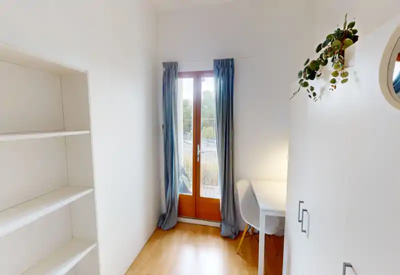 Compact bright living area with a glass door opening to a balcony, soft wood floors, a small desk and cozy curtains—ideal for a studio setup.