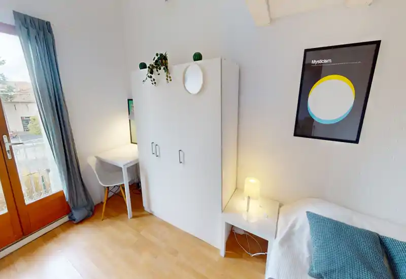 Small, bright bedroom with a comfortable single bed, bedside table with a lamp, a white wardrobe and a small desk beside the balcony door — neat, modern and well lit.