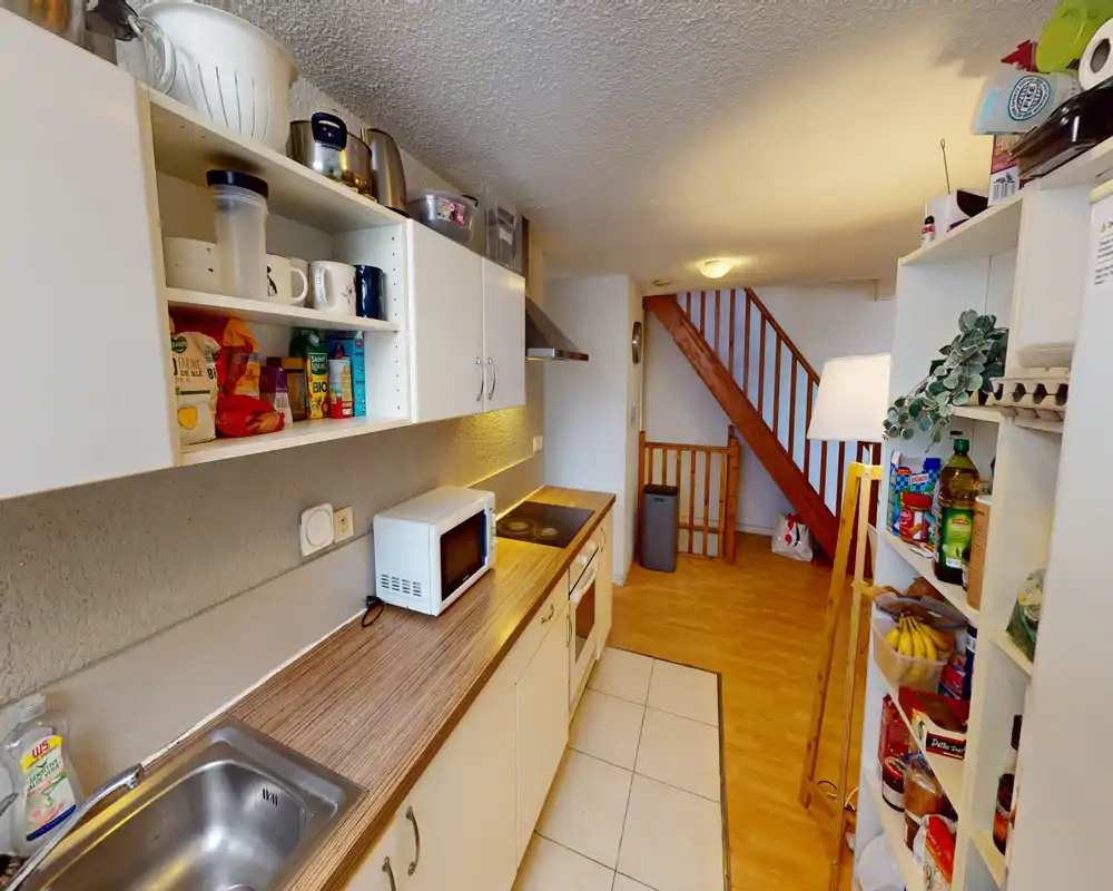 Narrow fitted kitchen with countertop appliances, open shelving and a pantry unit; warm wood flooring leads to a stairwell. Functional layout ideal for compact homes and holiday rentals.