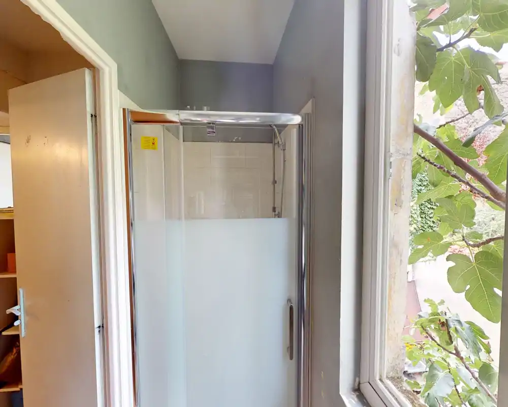 Compact bathroom view showing a corner shower cubicle with frosted glass door and a large window overlooking greenery.