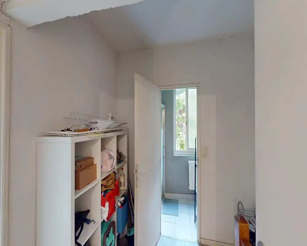 Small entrance or hallway with a storage shelving unit and an open door leading to a tiled room with a window. Neutral light and functional layout, suitable for storing coats and everyday items.