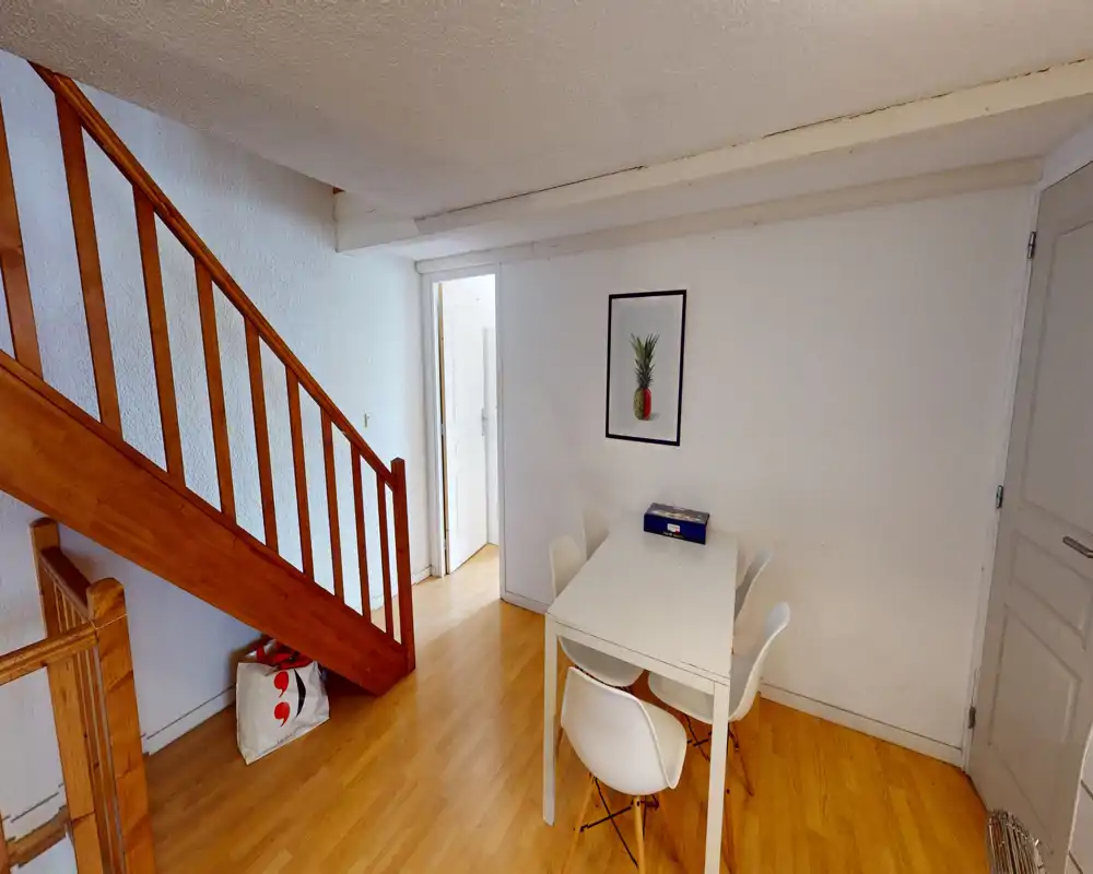 Compact dining area with a white table and four modern chairs next to a wooden staircase; clean minimalist styling and warm wooden floor.
