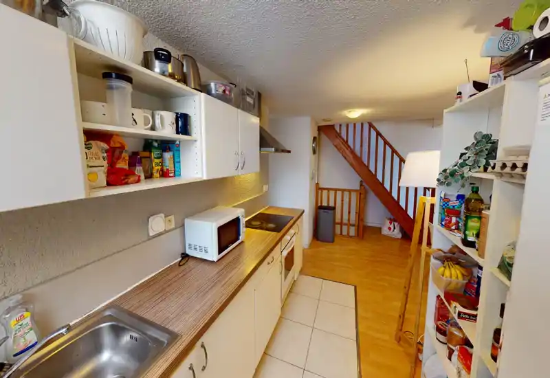 Cuisine étroite et équipée avec plan de travail, petits appareils et étagères ouvertes ; un sol en bois chaleureux mène à l'escalier. Agencement fonctionnel pour petits logements ou locations.