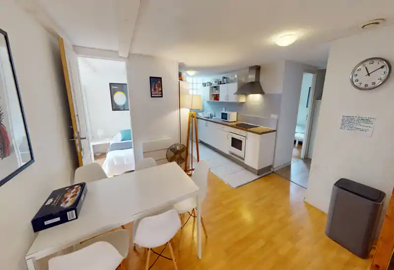 Bright open-plan living area with a dining table and visible kitchenette; modern white furniture and wooden floors create a clean, functional space ideal for short stays.