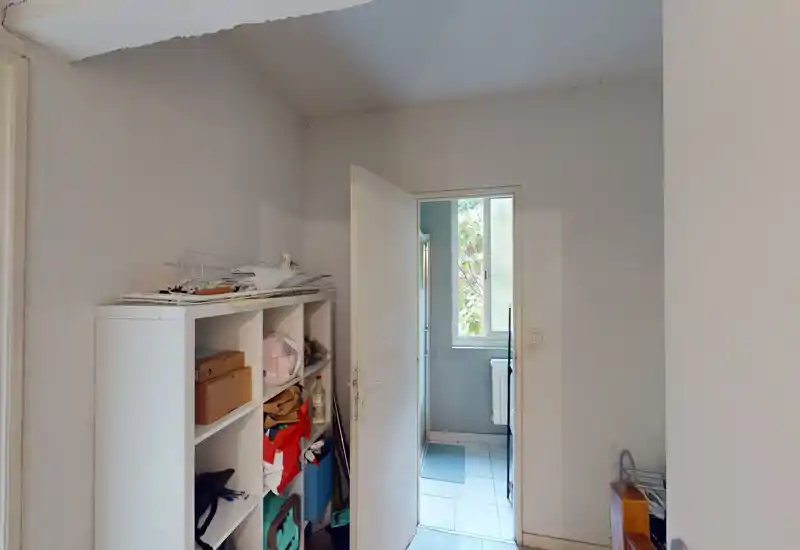 Petit hall d'entrée avec une étagère de rangement et une porte ouverte menant à une pièce carrelée avec une fenêtre. Lumière neutre et agencement fonctionnel, approprié pour ranger manteaux et effets du quotidien.
